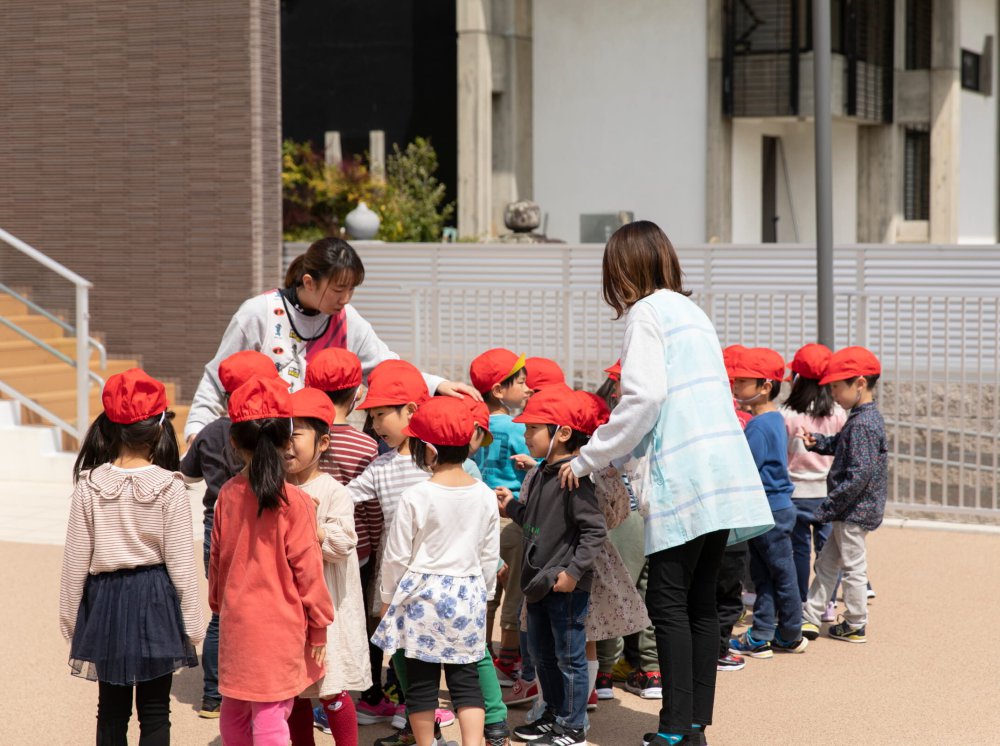 赤い帽子をかぶった園児たちを屋外で見守るスタッフ